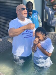 A man in a t-shirt stands next to a child in a water baptism ceremony.
